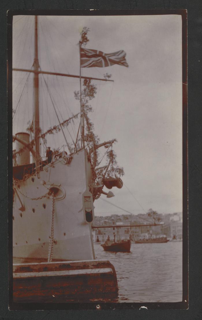 Prow of a British surface warship with Union Jack hoisted on jackstaff, itself also adorned with leaves/vegetal decorations